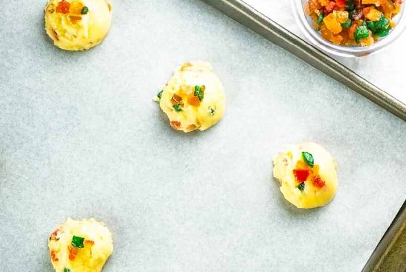 Fruitcake cookie dough on a baking sheet next to a bowl of candied fruit.