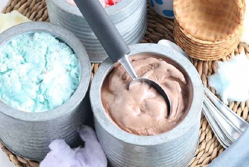 Three metal canisters filled with ice cream and a scoop with waffle bowls in the background.