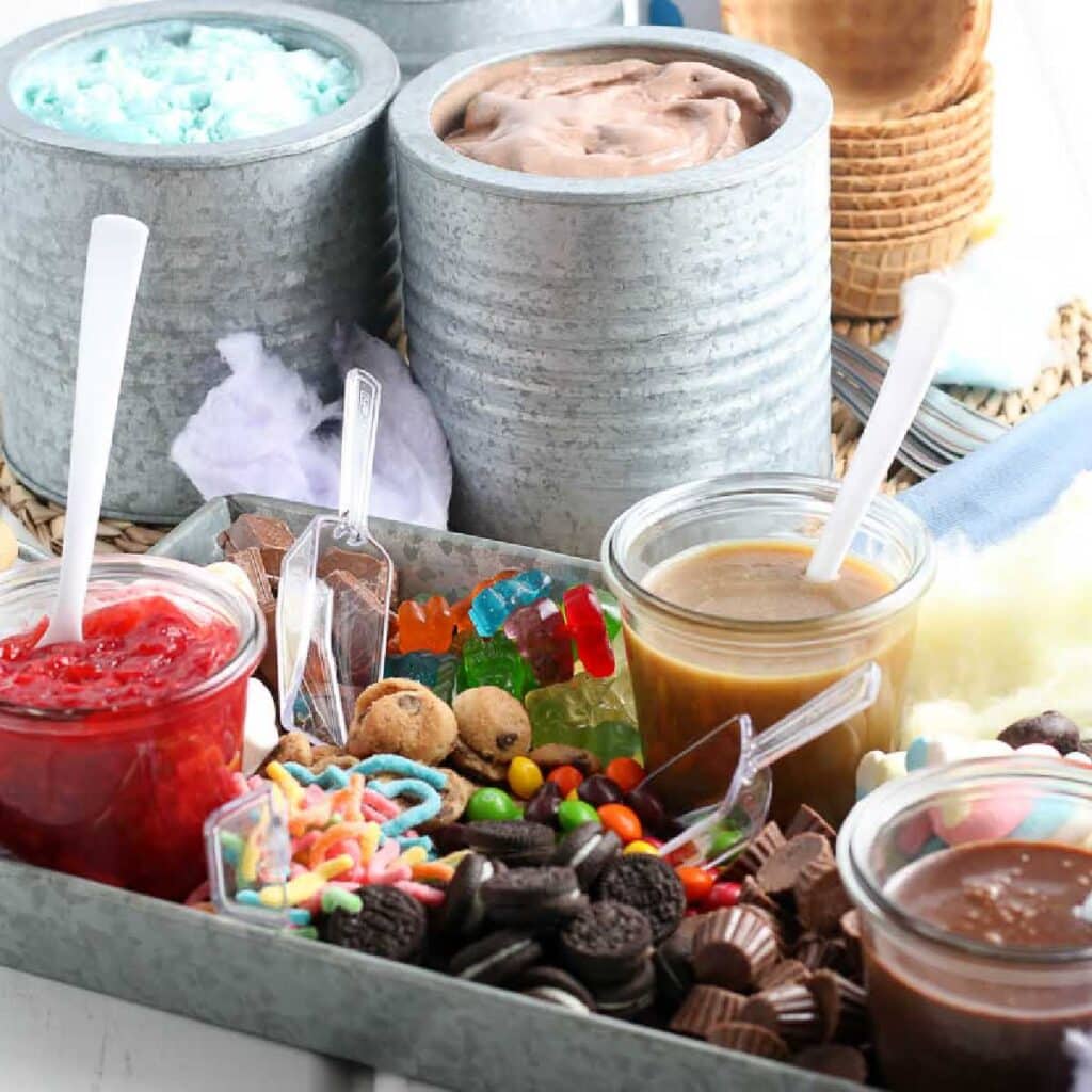 Metal serving tray filled with ice cream sauces and toppings with scoops and canisters of ice cream in the background.