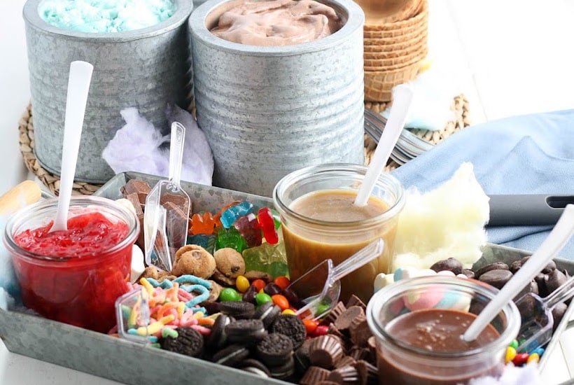 Metal tray filled with ice cream sauces and toppings with canisters of ice cream in the background.
