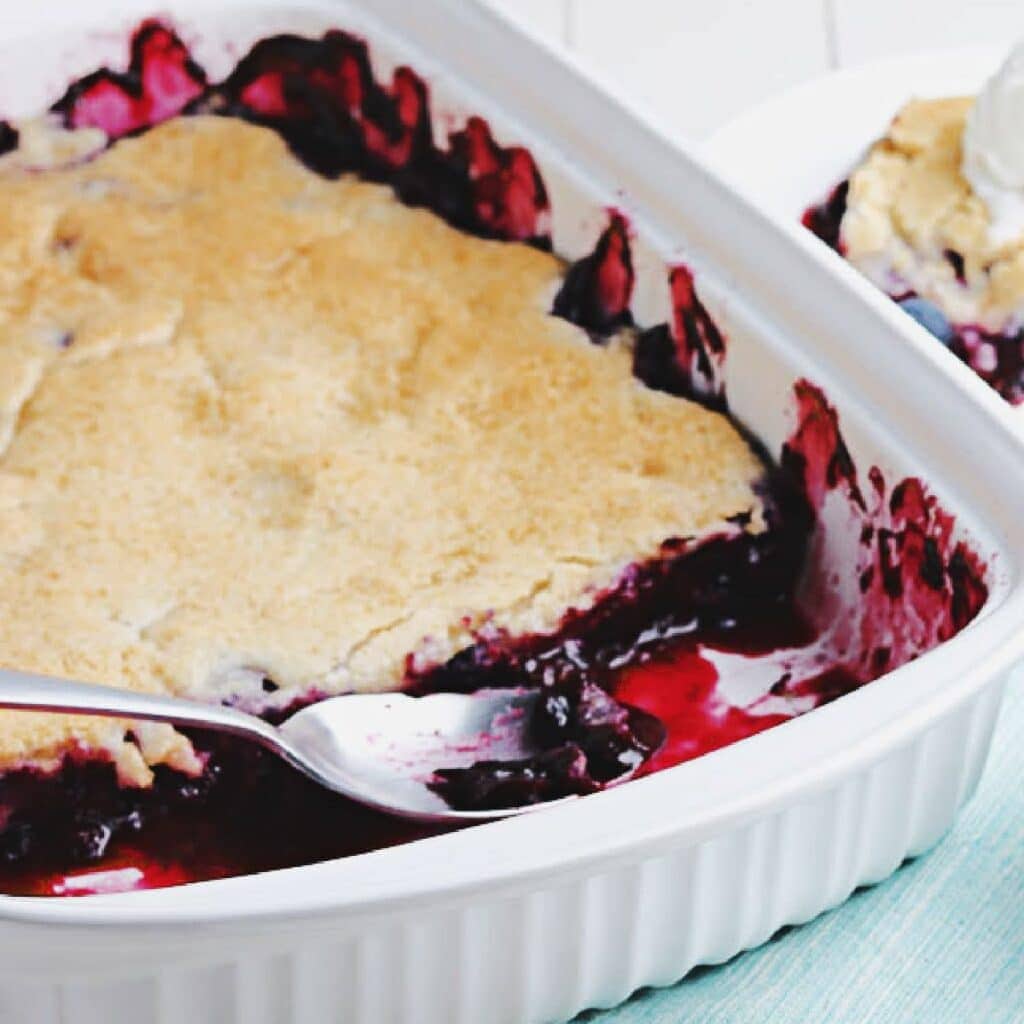 Old fashioned blueberry cobbler in a white casserole dish with a spoon inserted in the dish.