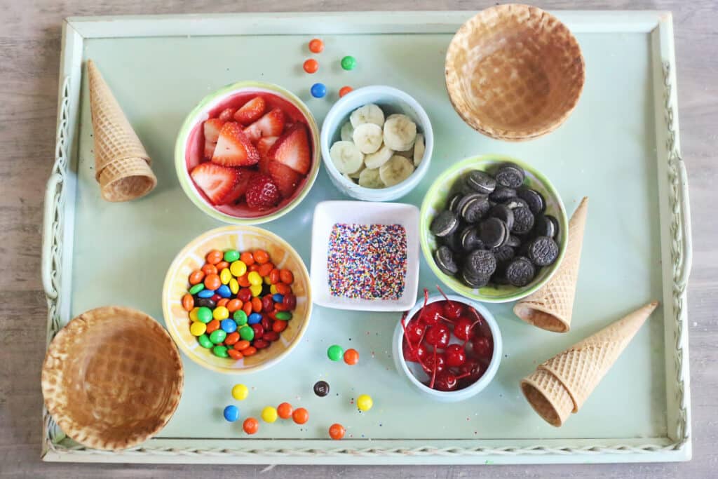 A tray filled with bowls of ice cream toppings and sugar cones and waffle bowls.