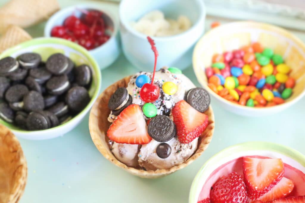 A waffle bowl filled with scoops of ice cream and surrounded by bowls of sundae toppings.