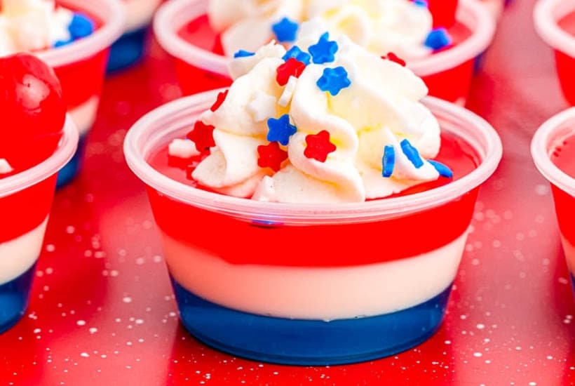 Layered patriotic jello shots with whipped cream and sprinkles on a red tray.