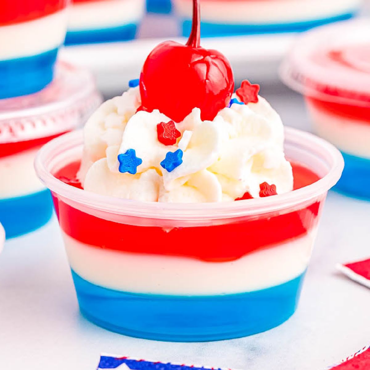 Closeup view of Red white and blue jello shot garnished with whipped cream, sprinkles and a cherry.