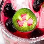 A glass of margarita on the rocks with blackberries and jalapeno slices.