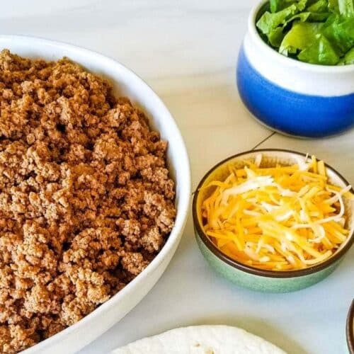 A bowl of taco meat with taco toppings nearby.