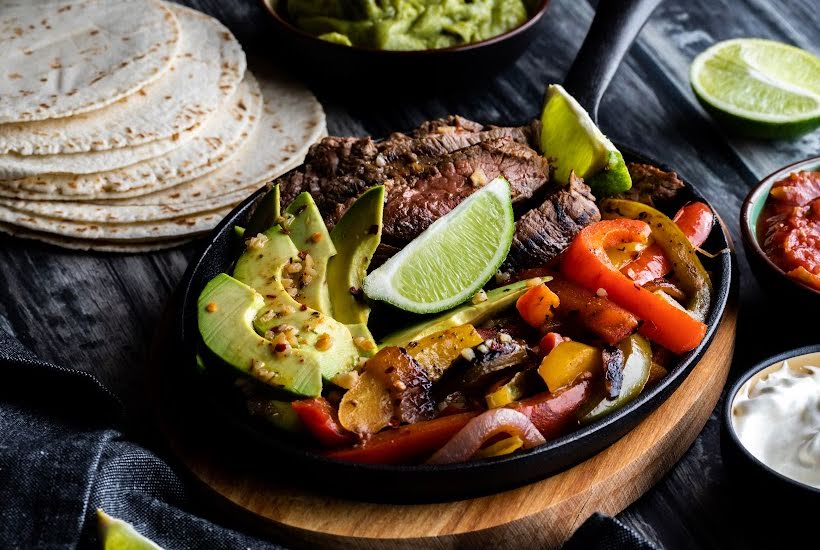 Skillet filled with steak, lime wedges and sliced vegetables with a stack of tortillas.