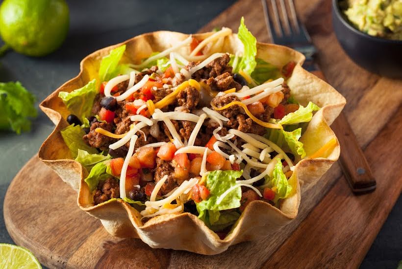 Taco salad in a tortilla bowl with ground beef, chopped tomatoes and shredded cheese.