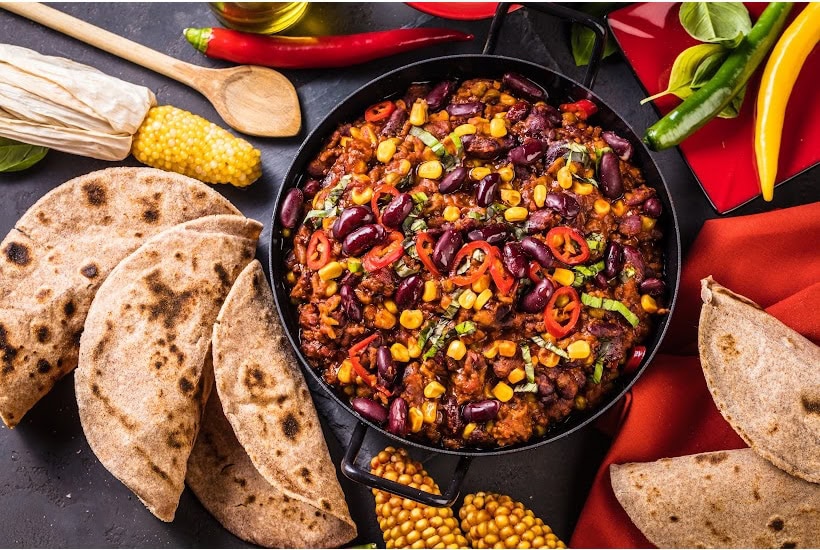 Skillet filled with taco meat, beans and corn surrounded by tortillas, peppers and corn on the cob.