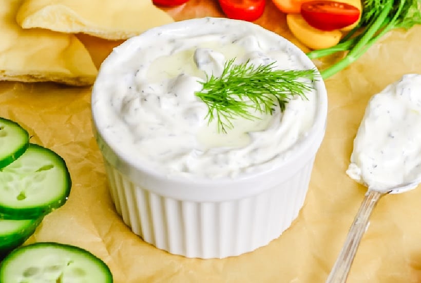 White ramekin bowl filled with creamy cucumber sauce garnished with fresh dill and surrounded by sliced veggies and pita bread.