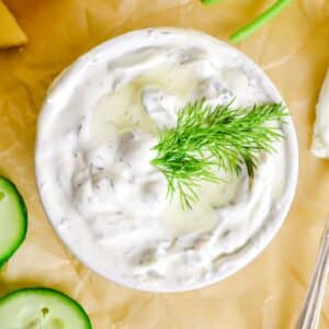 Bowl of creamy tzatziki dip with sliced cucumbers on parchment paper.