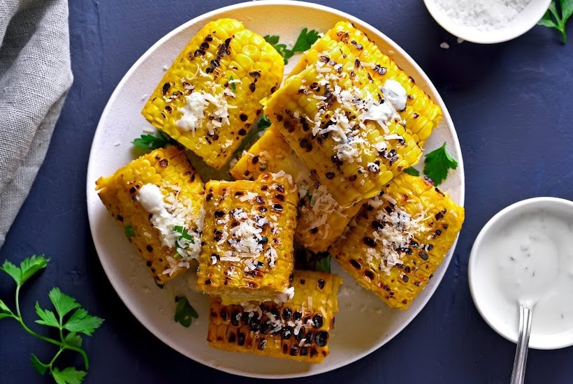 Mexican corn on the cob piled on a white plate with bowls of dipping sauces.