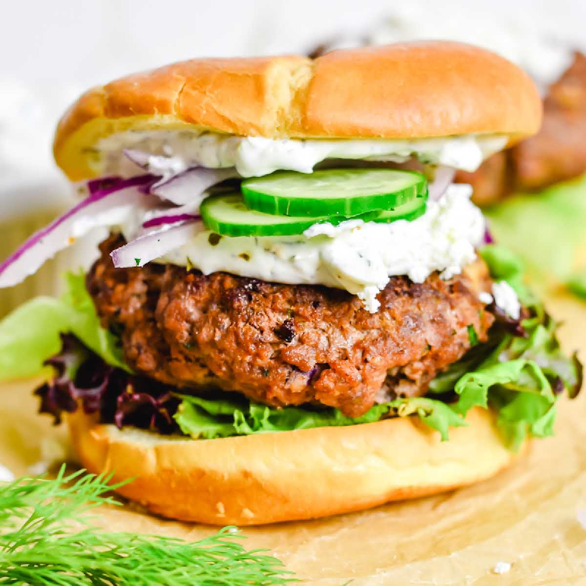 Closeup view of a Greek burger topped with feta cheese, sliced cucumbers, red onions and tzatziki sauce.