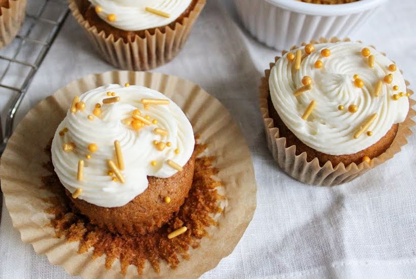 Frosted pumpkin cupcakes with brown butter frosting and sprinkles on a white tablecloth.