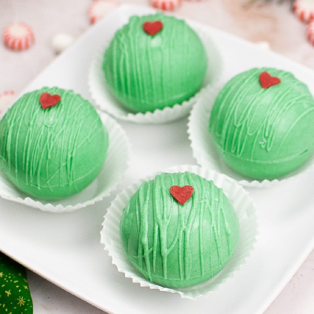 Four Grinch hot cocoa bombs on a square plate.