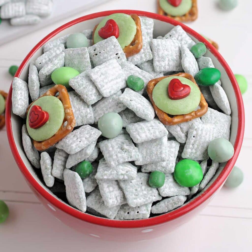 Closeup view of a bowl of Grinch Snack Mix with candies and pretzel bites with text overlay.